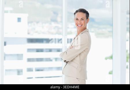Femme mi-adulte debout avec les bras croisés dans le bureau en costume beige près des fenêtres en verre, espace copie. Professionnel, affaires, leadership, confiant, bla Banque D'Images