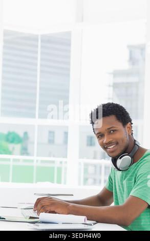 Homme afro-américain assis au bureau dans le bureau en utilisant des notes avec écouteurs et tablette, espace de copie. Individuel, lumière du jour, urbain, skyline, espace de travail, fo Banque D'Images