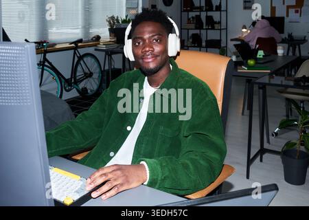 Portrait d'un jeune homme afro-américain travaillant à l'ordinateur de bureau tout en portant des écouteurs au bureau, représentant la productivité ciblée. Idéal pour le travail à distance, le support technique, le marketing de démarrage Banque D'Images