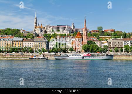 Une vue dégagée montre Buda Hill et le Danube à Budapest. Des bâtiments et des arbres bordent le front de mer. Les bateaux d'excursion reposent sur le quai. Banque D'Images