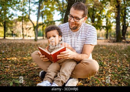 Dans un parc d'automne animé, un père partage un moment d'amour avec son enfant, lisant un livre ensemble entouré de feuilles colorées tombant. La chaleur de la famille est palpable. Banque D'Images