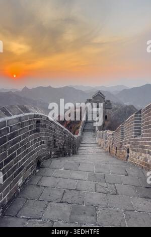 Vue verticale de la Grande Muraille de Chine au coucher du soleil. Le coucher du soleil brille sur d'anciens murs de pierre. Des escaliers mènent vers le haut à travers des pics montagneux accidentés. La lumière dorée peint le ciel dans des teintes ambre douces. Des tours lointaines se dressent fièrement au milieu de collines brumeuses Banque D'Images