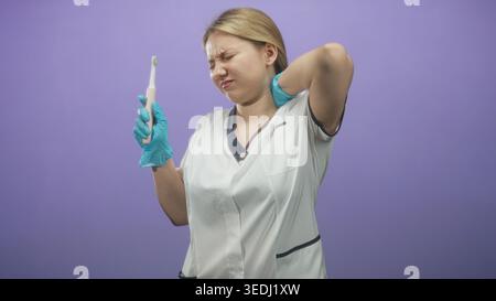 Femme dentiste portant un uniforme blanc et des gants bleus tenant une brosse à dents électrique, main sur le cou griffant dans le studio ; gêne. Banque D'Images