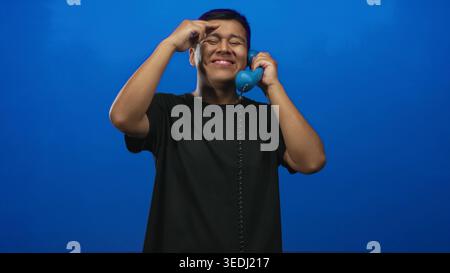 Jeune homme hispanique souriant tout en tenant le combiné téléphonique bleu dans le studio avec fond bleu vif ; joie décontractée. Banque D'Images