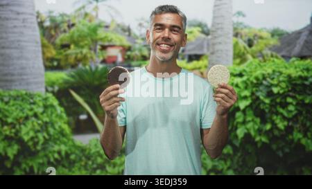 Homme tenant un gâteau ricecake et un biscuit au chocolat dans chaque main, langue dehors tout en dégustant une pâtisserie de maïs et des options de pesée dans une forêt de jardin de villégiature ; curio Banque D'Images