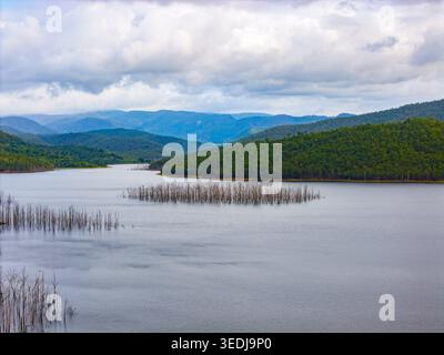 Un réservoir paisible avec des troncs d'arbres squelettiques s'élevant d'une eau calme sur fond de collines et de montagnes luxuriantes. Banque D'Images