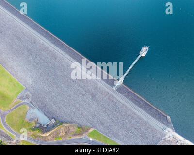 Tir par drone à grand angle capturant une structure massive de barrage en remblai de roche, un réservoir bleu tranquille et une tour d'admission technique, mettant l'accent sur l'ingénierie humaine Banque D'Images
