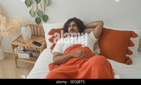 Homme allongé sur le lit avec une couverture orange serrant la poitrine et la main sur la gorge à côté de la table de chevet et smartphone dans le studio ; repos inconfortable. Banque D'Images