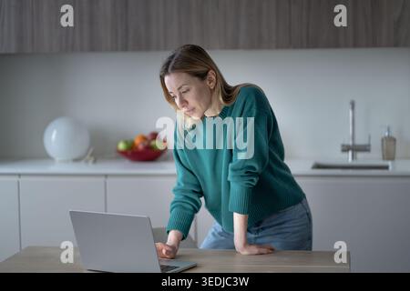 Femme d'âge moyen debout appuyée sur la table tout en regardant attentivement l'ordinateur portable dans la cuisine. Télétravail féminin ciblé ou navigation en ligne à la maison. Banque D'Images