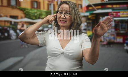 Femme hispanique senior pointe le doigt à la tempe portant des lunettes et le dessus blanc sur le marché de rue animé ; réflexion réfléchie. Banque D'Images