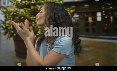 Jeune femme hispanique en chemise bleue souffle doucement baiser avec les deux mains sur la rue ensoleillée ; affection amour. Banque D'Images