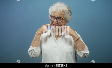 La femme senior fait des gestes avec les index sur ses joues puis pointe les deux index vers le haut, portant des lunettes colorées et un chemisier blanc en studio ; pla Banque D'Images