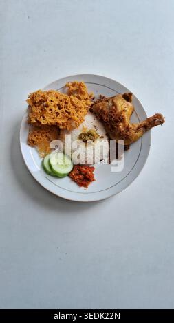 poulet frit traditionnel indonésien servi avec riz, tranches de concombre, sambal et craquelins croustillants, servi sur une assiette blanche sur une table blanche Banque D'Images