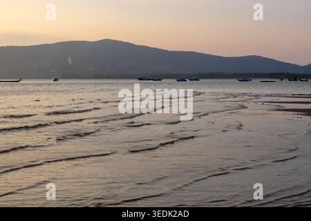 L'eau de mer tranquille ondule sur le sable sur la plage pendant le coucher du soleil avec des silhouettes de montagne et de bateau lointaines créant un paysage naturel paisible dans beautif Banque D'Images