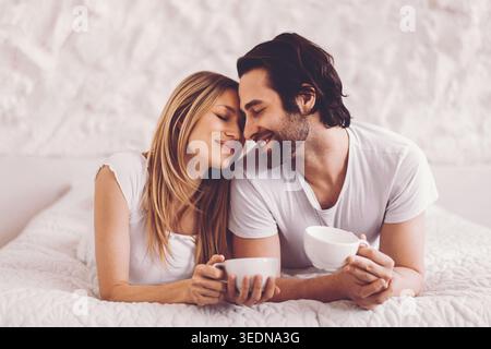 Couple profitant du matin ensemble tout en étant assis sur le lit avec des tasses à café dans les mains Banque D'Images