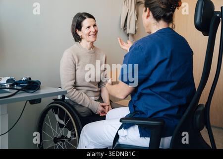 Femme mature souriante assise sur un fauteuil roulant écoutant le médecin féminin pendant l'examen médical dans la chambre d'hôpital Banque D'Images