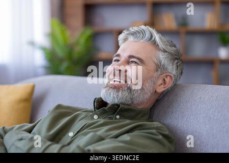 Homme mature avec les cheveux gris et la barbe assis sur un canapé gris à la maison, souriant avec satisfaction et se relaxant dans un salon calme et confortable moment de confort et de bien-être Banque D'Images