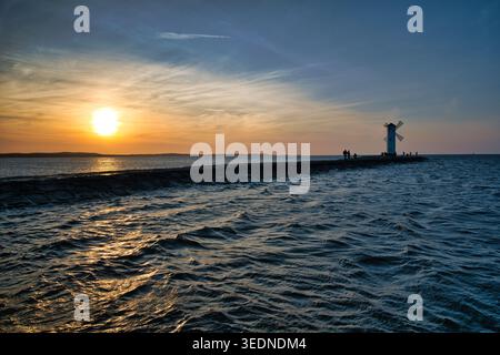 Phare de moulin à vent au coucher du soleil sur la jetée avec silhouettes sur Ocean Horizon - la ville balnéaire de Świnoujście. Banque D'Images
