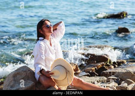 Femme assise sur les rochers tenant chapeau de paille par mer Banque D'Images