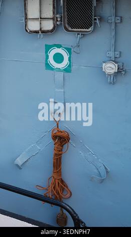 Plan vertical d'une station de sécurité maritime sur une coque bleue de navire avec un panneau de bouée de sauvetage, des crochets métalliques et une corde orange enroulée. Banque D'Images