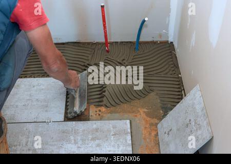 Le travailleur étale du mortier sur le sol tout en se préparant à installer des carreaux dans la salle de bain pendant les travaux de rénovation de la maison. Banque D'Images