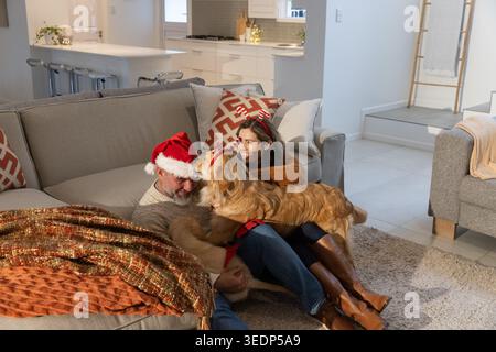 Homme senior portant chapeau de Père Noël et fille jouant à la maison avec Golden retriever portant harnais Banque D'Images