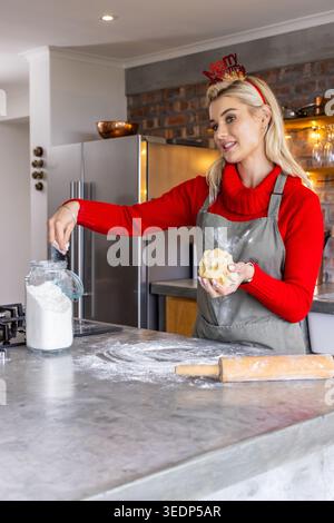 Femme portant la pâte à pétrir supérieure rouge tout en ramassant la farine du pot sur le comptoir, rouleau à pâtisserie Banque D'Images