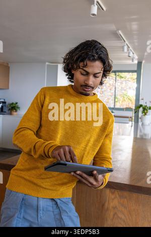Homme asiatique debout à l'îlot de cuisine tapant la tablette, portant un pull moutarde et un pantalon bleu clair Banque D'Images