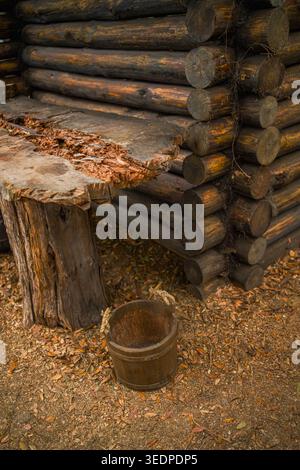 Une dalle de table en bois brut sculpté positionnée à côté d'un mur traditionnel de cabane en rondins avec un seau rouillé antique reposant sur des feuilles de chêne tombées sèches. Banque D'Images