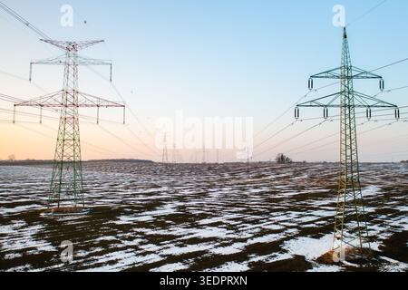 Des tours de transmission de puissance se dressent au-dessus d'un champ enneigé au crépuscule. L'horizon présente un doux dégradé de couleurs lorsque le soleil se couche, projetant une lumière douce sur le paysage hivernal. Banque D'Images