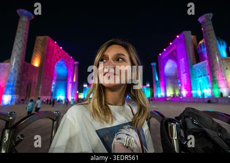 Une jolie touriste féminine se tient devant la place du Registan la nuit à Samarcande, en Ouzbékistan, illuminée par des lumières colorées. Banque D'Images