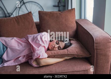 Une femme millénaire solitaire repose sur un canapé, ayant l'air ennuyée et déconnectée, éprouvant de la fatigue émotionnelle et une humeur faible tout en passant du temps seule à la maison Banque D'Images