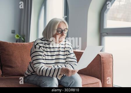 Femme âgée concentrée des années 60 assise sur le canapé dans le salon lisant la correspondance postale ou l'avis bancaire, femme mature concentrée des années 50 repos sur le canapé à la maison considérer le message dans la lettre de poste papier Banque D'Images