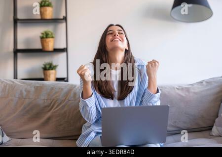 Une femme excitée est assise sur le canapé tenant un ordinateur portable et célèbre avec un geste joyeux oui après avoir reçu de bonnes nouvelles, comme une offre d'emploi de rêve ou une opportunité en ligne réussie Banque D'Images