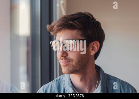 Positif rêveur beau jeune homme d'affaires dans des lunettes à la mode, regardant la fenêtre loin, pensant, rêvant, souriant, riant. Beau jeune homme modèle candide portrait Banque D'Images