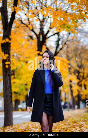 Jeune femme marchant en toute confiance sur un chemin couvert de feuilles mortes, portant un manteau noir élégant et un pull bleu, entourée d'arbres d'automne dorés vibrants Banque D'Images