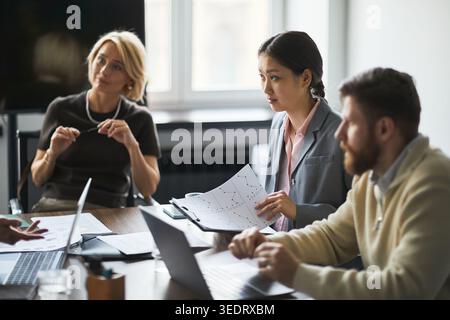 Jeune femme d'affaires asiatique présentant des graphiques financiers lors d'une réunion d'équipe avec des collègues utilisant des ordinateurs portables, discutant de stratégie et de planification. Idéal pour la communication d'entreprise ou le marketing sur le lieu de travail Banque D'Images