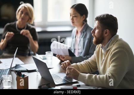 Homme d'affaires discutant de stratégie avec des collègues multiethniques lors de la réunion de bureau, en utilisant des ordinateurs portables et des documents. Idéal pour le travail d'équipe, le leadership, la planification, la communication d'entreprise Banque D'Images