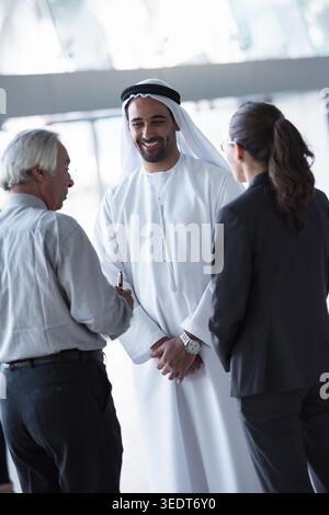 Un homme d'affaires émirati dans une kandura blanche et un ghutra sourit et discute avec deux collègues dans un bureau moderne lumineux, véhiculant professionnalisme, fierté culturelle et relations commerciales internationales amicales. Banque D'Images