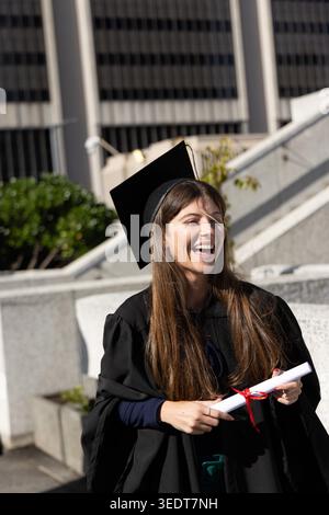 Femme portant mortarboard dans la robe posant sur plaza rire et tenant un diplôme avec ruban rouge. Diplômé, célébration, réalisation, certificat, campus, ensoleillé, extérieur Banque D'Images