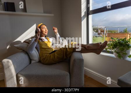 Femme asiatique s'inclinant dans un fauteuil à la maison portant une combinaison jaune écoutant avec un casque, téléphone sur les genoux. Sunlight, écouteurs, tablette, fenêtre, plante, cosy, détente Banque D'Images