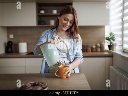 Une femme verse de l'eau chaude d'une bouilloire dans une tasse pour le thé. Elle se tient dans une cuisine avec des armoires en bois. Il y a des cookies sur la table. Banque D'Images