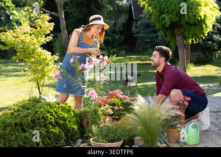 Bel homme excité admirant femme rousse alors qu'elle travaille dans le jardin, élaguant habilement des fleurs roses Banque D'Images