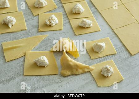 Carrés de pâte à pâtes avec garniture de fromage blanc et raviolis pliés sur une surface en bois. Banque D'Images