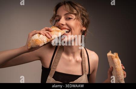 Jeune femme joyeuse dans un tablier tenant et mangeant de la baguette fraîche, appréciant le goût de la boulangerie et montrant l'appétit et la joie Banque D'Images