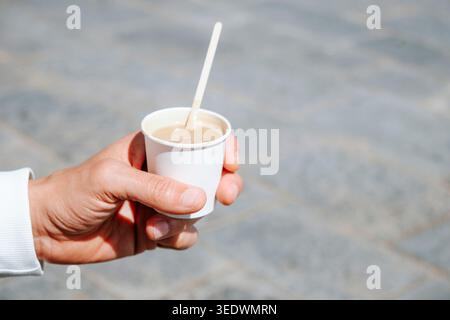 gros plan d'un homme tenant un cortado à emporter ou un expresso macchiato, un café avec une petite quantité de lait, servi dans une tasse de papier blanc, sur une urba ensoleillée Banque D'Images