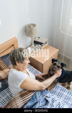 La femme âgée se réveille, caressant son chien assis à côté du lit dans une chambre lumineuse, commençant une nouvelle journée ensemble. La vie réelle d'une personne âgée Banque D'Images
