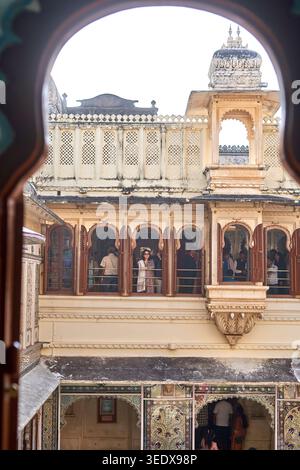 Vue à travers un cadre de fenêtre en bois en arc d'une cour de palais indien traditionnel, mettant en valeur une architecture rajastani à plusieurs étages avec des ornements Banque D'Images
