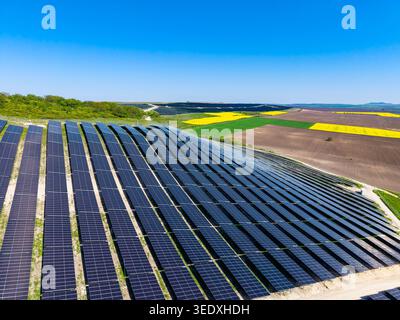 Les panneaux solaires couvrent une large zone sur les terres vallonnées. Les champs verts et jaunes s'étendent en arrière-plan. Le soleil brille dans un ciel clair. Cette configuration Banque D'Images