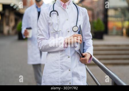 Travailleur de la santé portant une blouse de laboratoire blanche et un stéthoscope, tenant une planchette à pince et des dossiers tout en marchant avec un collègue à l'extérieur d'une clinique ou d'un hospita Banque D'Images